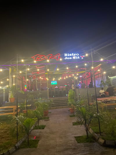 This is a nighttime, outdoor shot of a brightly lit bistro or restaurant called "Bistro Jos Cit". The sign for the bistro is prominently displayed in red neon letters with a stylized rose image above the "Jos Cit" text. String lights with yellow bulbs are draped across the scene, creating a festive atmosphere.
The bistro appears to have an open-air or semi-open-air seating area with a roof supported by a metal structure. Inside, there are glimpses of people seated at tables, and a television screen is visible in the background. The lighting is a mix of colorful neon signs, yellow string lights, and interior lighting, contributing to a vibrant and lively ambiance.
The foreground features lush green plants, possibly palm trees, in pots and growing alongside a concrete pathway. The ground appears wet, suggesting recent rain or damp conditions, and the time of day is clearly night due to the darkness of the sky and the reliance on artificial lighting. The overall mood is one of casual dining and evening entertainment. The location is identified as Rukuba Road, Nigeria.