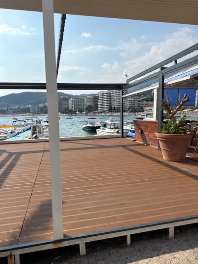 The image depicts a waterfront scene in Rafailovići, Montenegro, likely from a restaurant or cafe. In the foreground, a brown wooden deck extends across the lower portion of the frame, with a white vertical support post positioned off-center to the left. Beyond the deck, a body of water is visible, dotted with several white motorboats moored and at anchor. In the background, a coastal cityscape lines the shore, characterized by numerous multi-story buildings and a backdrop of lush green hills. The sky above is a clear blue with scattered white clouds, and a small bird is captured in flight against the sky. On the right side of the deck, a large terracotta pot holds a small tree with green foliage. The overall impression is a sunny, relaxed day at a coastal resort.
