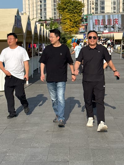 The image shows three men standing in a paved public space. The man at the top of the frame is wearing sunglasses, a black shirt, and black pants. He is holding a phone in his hand. The middle man is wearing glasses, a black shirt, and blue jeans. The man at the bottom is wearing glasses, a white shirt, and black pants. Behind the men are buildings, trees, and flags. A sign in the background indicates the location is the Shapingba District in China.