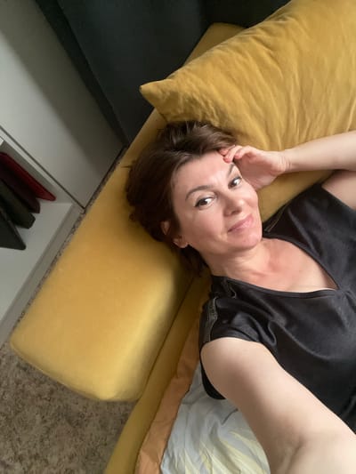 A woman lies on a yellow sofa, her head tilted back and resting on a cushion. She is wearing a black t-shirt with zippers on the shoulders and has her left arm bent, with her hand near her face. Her expression is relaxed, and she looks directly at the camera with a slight smile. The sofa is a vibrant yellow, and part of a dark gray sofa is visible to the left. In the background, a striped blanket or sheet is partially visible. Below the sofa, a white drawer unit is open, revealing a few items, including what appear to be small purses or wallets in red, dark green, and black. The flooring appears to be a speckled, light brown carpet. The lighting suggests it is daytime, possibly indoors. The scene conveys a sense of casual comfort and relaxation.