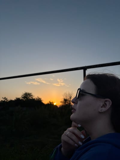 A profile view captures a young woman with dark hair, wearing glasses, against a backdrop of a setting sun. She has fair skin, an ear piercing, and manicured white nails. She wears a blue garment. Her hand is near her face. Behind her, a fence is visible against the gradient sky, and silhouetted trees and vegetation. The sun is positioned near the horizon. The location is Novopokrovskaya, Russia.