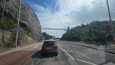 The image depicts a view from a vehicle driving on a road in Bristol, United Kingdom. The scene is dominated by a large, steep rock face on the left side of the road, with lush green trees and vegetation covering the hills in the background on the right. A suspension bridge, likely the Clifton Suspension Bridge, spans across the horizon. The sky is partly cloudy with a bright blue hue.
Several cars are visible on the road, including a red hatchback in the foreground with the license plate "WR74 CMY". Traffic lights are red, indicating a stop. A sign on a lamppost near the rock face reads "End of bus lane" in blue and "The Clifton Collection" in yellow with an arrow.
The time of day appears to be daytime, given the bright sunlight and clear sky, although there are scattered clouds. The weather seems pleasant, possibly a summer day. The overall atmosphere is that of a scenic drive through an urban landscape with natural geological features. There are no people clearly visible in the image, other than implied occupants of the vehicles. The image captures a typical driving scene in Bristol, highlighting the city's unique geography.
