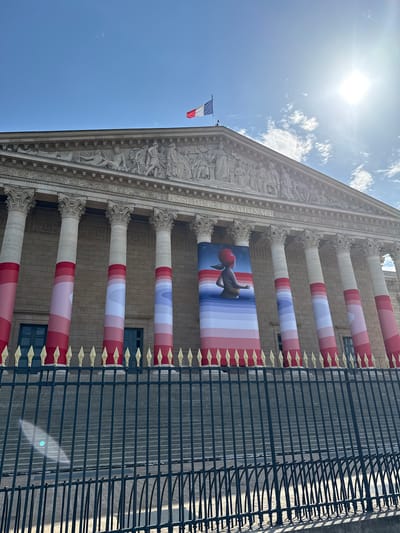 The image shows the exterior of the Palais Bourbon, the seat of the French National Assembly in Paris, France. The building features classical architecture with rows of large columns and detailed friezes. A French flag is visible flying from a pole on the left side of the building.
Several large banners are draped across the columns, displaying a pattern of red, white, and pink stripes. One banner in the center features a stylized cartoon character with a red head. The banners are likely part of a temporary exhibition or decoration. A black wrought-iron fence with gold finials runs along the front of the building, separating it from the viewer.
The sky is blue with some white clouds, and the sun is bright, casting shadows. The lighting suggests it is daytime, likely late morning or early afternoon, given the strong sunlight and clear sky. The overall scene conveys a sense of grandeur and public space. The text "ASSEMBLÉE NATIONALE" is partially visible on the building's facade.
