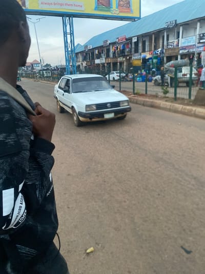A medium-shot, eye-level, outdoor, daylight scene captures a street in Bukuru, Nigeria. In the foreground, a person's left shoulder and back are visible, draped with a black jacket featuring a white pattern and text. The person is looking forward, with their left hand grasping the jacket's strap.
Dominating the mid-ground is a white Volkswagen car, its front facing the viewer's left. The car is on a paved road. Beyond the road, a row of two-story buildings forms a marketplace. These buildings have light blue roofs and balconies. Numerous stalls and shops are visible on the ground floor, displaying various goods, including clothing, plastic containers, and what appears to be food items. Green fencing separates the road from the marketplace.
In the background, a large blue metal structure supports a billboard with yellow and white text. The sky is overcast. The overall atmosphere suggests a typical urban environment in Nigeria.