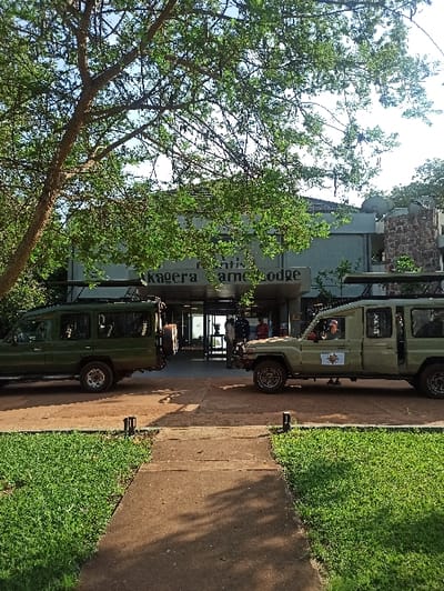 Two safari vehicles park in front of the Rntis Akagera Game Lodge. The lodge's signage is visible above the vehicles. A tree dominates the left foreground. A paved walkway separates the vehicles from a well-maintained grassy area. The building appears to be multi-storied, with stone elements visible on the top levels. The sky is partly visible through the tree's branches.
