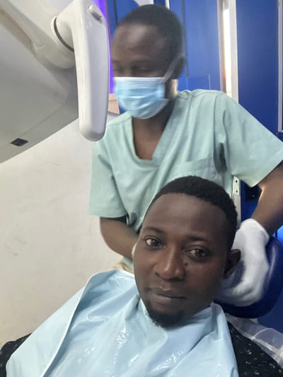 Two Black individuals are depicted in what appears to be a medical setting, possibly a dental clinic, in Rido, Nigeria. The foreground is occupied by a man wearing a light blue dental bib, with a faint beard and dark hair. He is looking directly at the camera with a neutral expression. Behind him, another individual, also Black, is partially visible, wearing a light green medical scrub and a light blue surgical mask. This person is standing and appears to be a medical professional, as indicated by the gloves on their hands and the light blue mask. They are positioned behind the seated individual, with one gloved hand resting on the back of the dental chair or the other person's shoulder. The background shows a bright white wall on the left, a white medical apparatus with a handle in the upper left quadrant, and a blue wall or partition on the right. A fluorescent light fixture is also visible on the right side. The overall lighting is bright and consistent with an indoor medical environment.