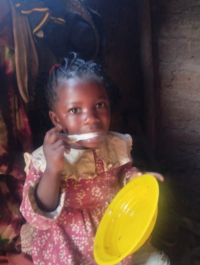 A young, dark-skinned girl holds a yellow plate of food in her right hand and a spoonful in her mouth. She is adorned in a long-sleeved dress, which has a white floral pattern on red with a frilly white collar. She has braided hair and is looking at the camera. The backdrop is a dimly lit room with clothes or fabric hanging in the upper left corner. A wooden beam is visible on the right side of the frame.