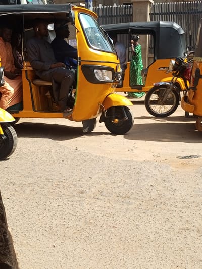 The image depicts a row of yellow auto rickshaws, locally known as kekes, on a paved street in Maiburiji, Nigeria. In the foreground, one keke is occupied by four passengers and a driver. The driver wears a dark shirt, and one passenger wears an earpiece. Another keke is visible in the mid-ground, with a passenger in green patterned clothing. Metal gates are visible in the background. The foreground shows details of the paved road.