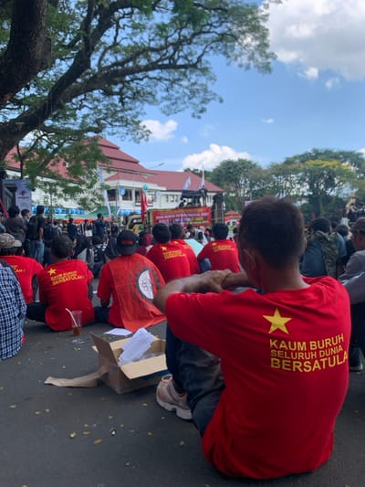 The image shows a group of people sitting on the ground in front of a building in Malang, Indonesia, located at coordinates -7.977 latitude, 112.634 longitude. They are all wearing red shirts with yellow stars and slogans in Indonesian, including "KAUM BURUH SELURUH DUNIA BERSATULA," which translates to "Laborers of the Whole World Unite." Some are wearing hats, and some are holding signs. There is a tree in the background. There is a large banner in the background that says, "HARIBO BURUH INTERNASIONAL SE-ASIA - 1 NO - 8 TAMAN 2023." The individuals are gathered around a cardboard box and are seated on a paved area. The scene is likely a labor protest or rally. It appears to be a peaceful gathering.