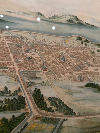 The image shows an antique map of Mexico City. It depicts a densely packed urban area characterized by closely arranged buildings, waterways, and roads. The river runs alongside the city's border, with a clear difference between the developed area and the surrounding landscapes. The map indicates areas such as parks or gardens, with distinct details. In the upper right corner of the image, the inscription "Agua de S. Fo yoo arces" can be observed.