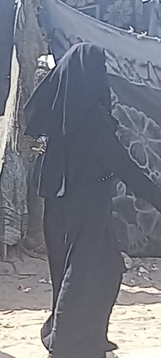 An outdoor scene in Khan Yunis, Palestinian Territory, shows a single individual, dressed in full black attire including a head covering, standing or walking. The person's face is not visible. Small light-colored embellishments are arranged vertically on the upper right side of the garment, and a white tag or fabric piece hangs from the left side. To the left of the individual, an irregular, light-colored structure or wall is visible. To the right, a large dark fabric panel, possibly a tarp or blanket, with a distinct light-colored floral or circular pattern, is draped vertically. The ground beneath the person is uneven, appearing to be dirt or sand, with scattered fragments of debris, including reddish-orange and white pieces.