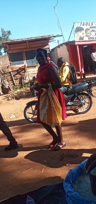 The image shows a woman in a red dress standing next to a motorcycle in Lwandeti, Kenya, located at latitude 0.563 and longitude 34.806. The woman is holding a yellow and white bag. Another man, wearing a yellow shirt and a black backpack, is sitting on the motorcycle. The motorcycle is parked in front of a barber shop. There are wooden buildings on the left side of the image. The ground is dirt. The woman's shadow is cast on the ground. There is a partially visible person in the foreground wearing a blue jacket. The sky is blue and cloudless.