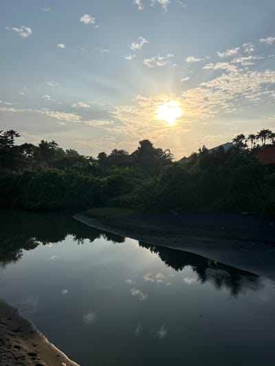 This vertical, outdoor, eye-level shot captures a serene water scene at sunset in Mengwi, Indonesia. The foreground features dark sand and the calm, dark water of a river or bay, reflecting the sky and surrounding trees. The middle ground is dominated by lush, dense green foliage, creating a thick wall of vegetation that lines the water's edge. Peeking through the trees are glimpses of buildings with terracotta roofs and silhouettes of palm trees against the sky. The sky above is a pale blue, transitioning to a soft yellow near the horizon. Sunlight beams dramatically through scattered, wispy white clouds, with the bright sun partially obscured by some of these clouds.