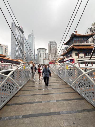 A slightly upward-angled, rotated shot captures a modern bridge in Yuzhong District, China, against a backdrop of skyscrapers and traditional Chinese architecture.
Several people are walking on the bridge. The bridge has a gray metal railing with circular cutouts. Wires from above stretch into the sky.
Traditional Chinese buildings with tiered roofs and decorative eaves are visible on the left, contrasting with modern high-rise buildings in the background. The sky is overcast. The bridge's surface is paved with rectangular tiles.