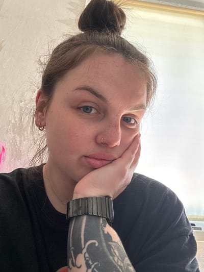A close-up selfie of a young woman with fair skin, light brown hair pulled back in a bun, and blue eyes. Her left hand is resting against her cheek. She has freckles, a nose piercing, and is wearing earrings. A black smart watch is visible on her left wrist, with part of a tattoo visible on her arm. She is wearing a black t-shirt and a silver necklace. The background is light and indistinct. There is no information to determine the location.