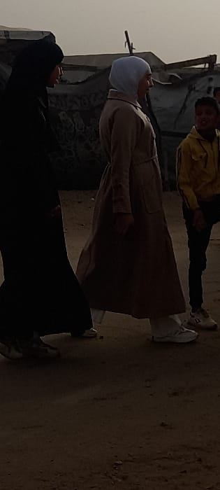 The image depicts an outdoor scene featuring three individuals on an unpaved, dusty ground. The overall lighting suggests it is daytime, possibly overcast or hazy, as there are no harsh shadows.
In the foreground, on the left side, stands a person wearing a long black garment, likely an abaya, and a black head covering that conceals their face, possibly a niqab. They are standing with their body angled towards the right, looking slightly upwards towards the person in the middle. They are wearing light-colored footwear.
In the center of the frame, another person is standing, dressed in a light brown or beige long coat or dress, reminiscent of an abaya, and a light blue or white head covering (hijab). This individual is facing slightly towards the left side of the frame (relative to their upright position), with their face visible in profile. They are wearing white sneakers.
Further in the background, towards the right, a young person, possibly a child or teenager, is visible. They are wearing a bright yellow long-sleeved top and dark trousers. This person is looking generally towards the viewer, with a slight smile on their face. Their hands are down at their sides, and they are wearing dark shoes.
The background consists of rough, possibly dilapidated structures or walls made of earth or stone, with visible textures and some debris. The sky above is a uniform light grey or tan, blending into the distant structures. The ground is uneven dirt or sand, with small rocks and irregularities. The individuals appear to be standing and observing, possibly engaged in a casual interaction or simply present in the outdoor environment. There is no visible text in the image.