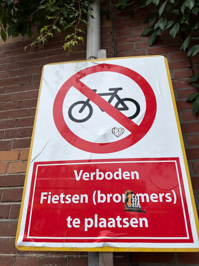 A Dutch prohibition sign is displayed on a brick wall, partially obscured by green foliage. The sign is yellow-bordered and features a white background with a prominent red circle and slash symbol overlaying a black silhouette of a bicycle, indicating that bicycles are not allowed. Below this graphic, a red rectangular section contains white text. The top line reads "Verboden" (Forbidden). The second line states "Fietsen (brommers)" (Bicycles (mopeds)), with a small sticker of a stadium and the text "1 CL STA Ajax Amsterdam" placed over the word "brommers". The third line reads "te plaatsen" (to place). The sign is attached to a light-colored pole. The background consists of a reddish-brown brick wall.