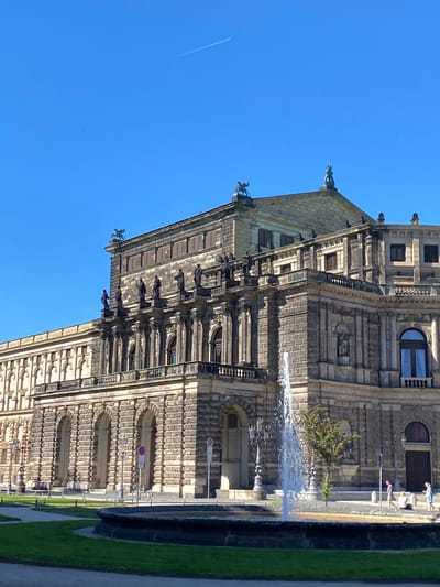 The image captures the Saxon State Chancellery in Dresden, Germany, under a clear blue sky. The building is constructed from light brown sandstone. There are statues of gargoyles and human figures on the building. A fountain is visible in front of the structure. The area surrounding the building is landscaped with green grass. A few people are visible.