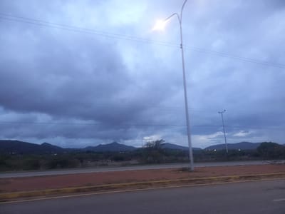 This image captures a roadside view of a landscape with mountains in the distance under a cloudy sky. A tall, slender streetlight with a bright light illuminates the scene from the right side of the frame. Multiple power lines stretch horizontally across the sky. In the foreground, a paved road with yellow and reddish-brown markings runs horizontally. Beyond the road, there is a strip of reddish-brown earth, followed by a dark area of vegetation and then the distant, dark silhouette of rolling hills and mountains. Several small, bright lights are scattered across the foothills and slopes of the mountains, suggesting the presence of settlements or structures. The overall atmosphere is overcast and somewhat dim, indicative of either early morning, late evening, or stormy weather. The scene does not contain any visible people or vehicles. The image appears to be taken from a moving vehicle.