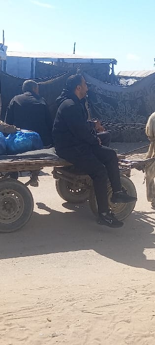 Two men in dark clothing are working on the underside of a horse-drawn carriage. The carriage has four wheels and is parked on a sandy surface. One man is lying on his back beneath the carriage, apparently making repairs. Blue tarp or material is visible near the carriage. The area appears to be an outdoor location, possibly a roadside or rural setting.