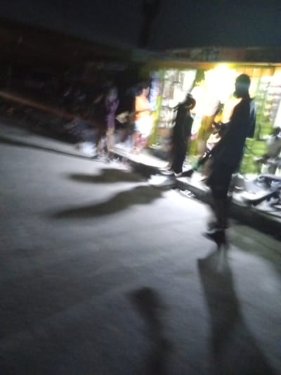 A poorly lit street scene in Ado, Nigeria, at night. A group of people are standing in front of a brightly lit storefront with items displayed in the window. Shadows of the people are cast on the gray pavement. The scene is blurred.