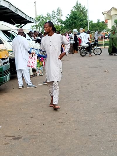 In Gombe, Nigeria, a man in a light-colored traditional outfit walks across a paved road. He carries a blue box with various items and a patterned shopping bag. Several white vans are parked behind him, and other individuals are visible in the background, some near the vehicles and others further away. A person rides a motorcycle to the right of the frame, and a woman in a green hijab is also present in the background. The scene appears to be an outdoor marketplace or transport hub, with buildings and trees in the distance. The lighting suggests it is daytime.