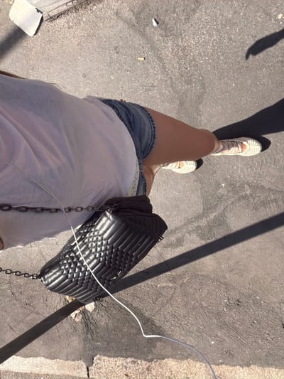 A person is standing on a pavement, casting a shadow. They are wearing a white t-shirt, denim shorts, and white sneakers with white laces. A black quilted handbag with a chain strap is slung across their body. A white cable is trailing from the handbag. In the background, there is a concrete structure and some scattered cigarette butts on the ground. The scene appears to be outdoors in an urban environment.