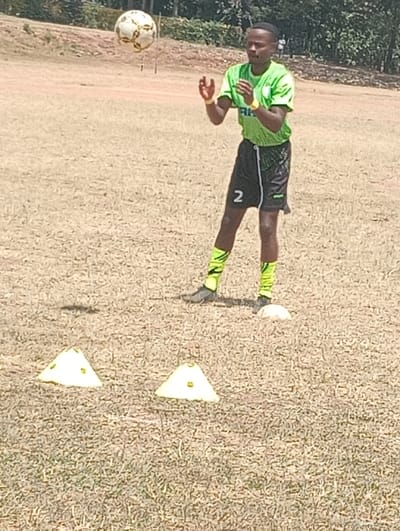A young man wearing a lime green soccer jersey with black shorts and black and white socks is practicing soccer on a grassy field. He has his hands up as he watches a soccer ball arc through the air. The ball is a white and black leather design. The man is wearing a yellow bracelet on his right wrist. There are yellow triangular cones on the ground. The ground is covered in dry, brown grass. The location is Kipkarren, Kenya, at coordinates (latitude: 0.596, longitude: 34.996).