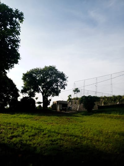 The image depicts a sunlit grassy field in Thalang, Laos, with several trees casting shadows across the foreground. In the midground, there is a building near a stone wall and a wire fence. A motorcycle is parked near the building. The sky is pale blue with some clouds. The scene is outdoors, and the environment appears natural and somewhat unkempt. There are no people visible in the image.