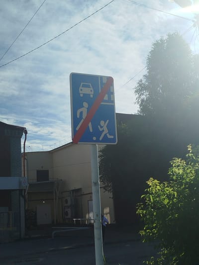 The image shows a street scene in Zavodoukovsk, Russia. In the foreground, a vertical blue traffic sign with a red diagonal line indicates the end of a restriction for vehicles and pedestrians. The sign depicts a white car at the top and two white figures, one adult and one child, walking below. The sign is mounted on a white pole. Behind the sign, a light-colored building with a dark entrance is visible, along with a portion of a brick building to the left. Overhead, power lines stretch across a cloudy sky, and a large tree with green foliage is present on the right side of the frame, partially obscuring the building. The lighting suggests it is daytime, with sunlight creating some lens flare. A person is partially visible on the far left, near the brick building.