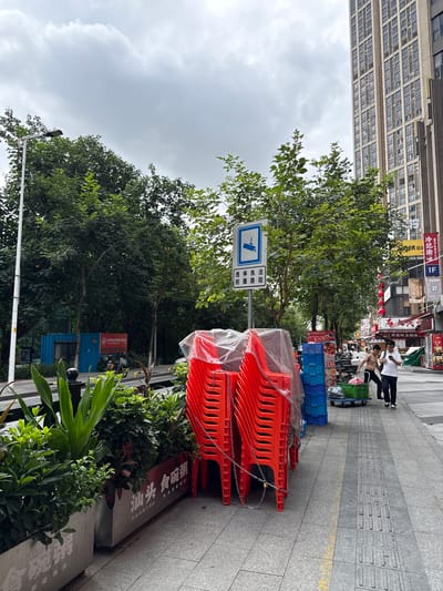 A street scene in Haizhu District, China, on a cloudy day. In the foreground, a stack of red plastic chairs is covered with a translucent plastic tarp and secured with chains. To the right of the chairs are stacks of blue plastic crates and a green cart laden with goods, being pushed by two men. The man on the left, shirtless, is looking at his phone, while the man on the right, wearing a white t-shirt and black pants, is also looking at his phone. They are walking on a sidewalk with textured paving for the visually impaired. Behind the chairs and crates, a planter box filled with lush green plants runs along the sidewalk. A street sign indicating surveillance is visible to the left of the chairs. Further down the street, there are shops with awnings, and pedestrians. A tall, modern building with many windows dominates the right side of the background. The street is paved, and there are trees lining the sidewalk. A blue shipping container is visible on the left side of the image.