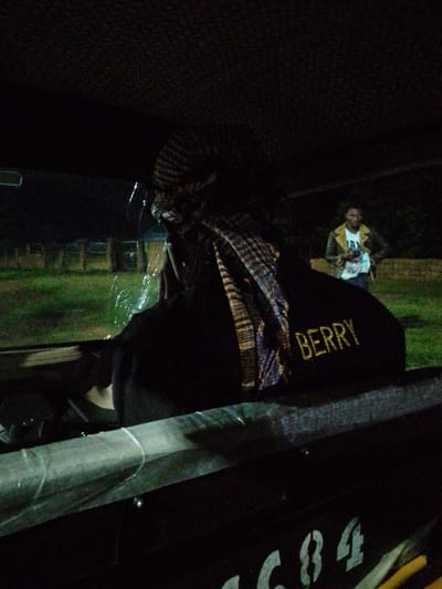 The image shows a dark, possibly nighttime scene, with the subjects inside a vehicle. The primary subject is a person from behind, wearing a headscarf and a black shirt. The word "BERRY" is visible in yellow lettering on the back of the shirt. Through the vehicle's window, a second person is visible standing outside. This person is wearing a white t-shirt with some lettering and a brown jacket. They are holding a phone. The setting appears to be outdoors, with grass and some structures visible in the background. The lighting suggests it is either late evening or nighttime, and the grass appears green, hinting at either a recently watered area or the presence of artificial lighting. There are no obvious weather cues, but the overall atmosphere is dark and somewhat grainy, typical of low-light photography. The image composition focuses on the interior of the vehicle and the person in the foreground, with the person outside being secondary.