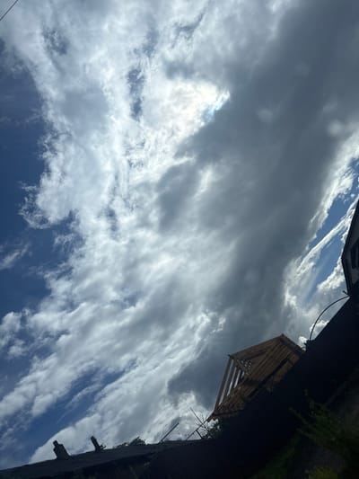 The image shows a view looking upwards towards a dramatic sky filled with white and grey clouds, interspersed with patches of blue. The sun is partially obscured by thicker clouds, creating bright highlights and darker shadows. In the foreground, a dark silhouette of rooftops and vegetation is visible. On the right side, a portion of a building with windows and a railing is seen. A wooden structure, possibly a framed roof or scaffolding, is positioned at an angle on the right side of the image, against the backdrop of the sky. The scene appears to be outdoors in an urban or semi-urban environment, potentially in Капыльскі сельскі Савет, Belarus, given the context. There are no people visible in the image, and no discernible actions or interactions are taking place. The environment is characterized by a mix of man-made structures and natural elements (sky and vegetation).