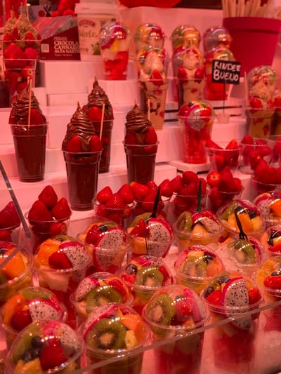 This is a medium-shot, eye-level, indoor photograph of a food stall displaying a variety of fresh fruit and chocolate-covered strawberry desserts, likely in Barcelona, Spain. The stall is brightly lit with pinkish-red lighting, giving the scene a vibrant and warm atmosphere.

In the foreground, the bottom half of the image is filled with numerous clear plastic cups containing meticulously arranged fruit salads and chocolate-covered strawberries. The fruit salads feature a colorful mix of chopped fruits such as dragon fruit (pitaya), kiwi, mango, strawberries, blueberries, and possibly pineapple or papaya. Some cups are topped with whole strawberries or pieces of dragon fruit. The chocolate-covered strawberries are presented in clear plastic cups, with the chocolate swirled decoratively over fresh strawberries.

In the middle ground, several rows of these fruit and dessert cups are displayed on tiered white shelves. Behind these, higher up, are more cups, including some filled with what appears to be yogurt or ice cream topped with fruit. A small black sign with white lettering reading "KINDER BUENO" is visible among the desserts. There are also containers of what looks like chocolate or caramel sauce.

In the background, the stall continues with more displays. A large, partially visible pink pot or container is in the upper right corner, and beyond that, more products are stocked. The overall impression is of a well-organized and abundant food display, possibly at a market or specialized dessert shop. There are no people visible in the image.