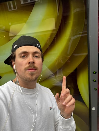 A male is centered in the image wearing a white sweater with an embroidered emblem on the upper right. He is wearing a black baseball cap and white wired earphones. He is pointing with his right hand and has tattoos on his wrist and neck. The background is a blurred image of yellow bananas. The top of the image shows a gray frame with green Monster Energy logos. This is a bus stop advertisement in London, United Kingdom.