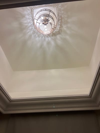 The image shows a close-up, low-angle shot of a decorative tiered chandelier hanging from a white recessed ceiling. The chandelier features multiple circular tiers with intricate metalwork, casting a detailed shadow pattern on the ceiling. The ceiling itself has a stepped design, with molding visible along the edges. The lighting is warm, emanating from the bulbs within the chandelier. The overall environment appears to be indoors, possibly in a hotel lobby or a well-appointed residential space. There are no people or animals visible in the image. The location is not identifiable as Sharm El Sheikh, Egypt, based on the visual information provided.