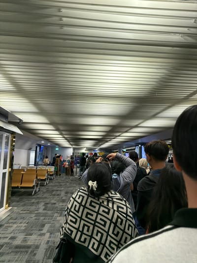 The image depicts a queue of people inside an airport terminal, likely at Suvarnabhumi Airport in Bangkok, Thailand, given the context of travel. The ceiling features a distinctive ribbed, metallic design, a common architectural element in modern airports. Fluorescent lighting casts long shadows across the ceiling and down the hallway. In the foreground, the back of a person wearing a patterned shawl is visible. Further down the queue, various individuals can be seen with luggage, suggesting they are either arriving or departing. The environment appears orderly, with seating areas visible to the left and information displays or signs further down the terminal. The overall scene conveys a sense of transit and waiting, characteristic of an airport setting.