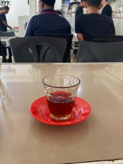 The image depicts a glass of tea on a red saucer, placed on a light brown table. In the background, several people are visible, some seated at tables and others standing. The setting appears to be an indoor establishment, possibly a cafe or restaurant, with white walls and shelves in the background. One of the individuals in the background is wearing a dark blue shirt and a black cap, and another is wearing a black t-shirt. The atmosphere suggests a casual gathering or meal. The scene is likely located in Lhokseumawe, Indonesia, a city known for its culinary scene and places where people gather for refreshments.