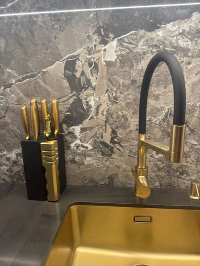 The image depicts a modern kitchen sink area with a prominent gold-colored sink and faucet. The faucet has a black hose and a gold-toned base and handle. Next to the sink, on a dark countertop, sits a black knife block holding several gold-handled knives. The backsplash is made of dark marble with white veining. The scene appears to be indoors, likely in a residential kitchen. There are no people visible in the image. The location is not specified to be in Sochi, Russia; this is an assumption.