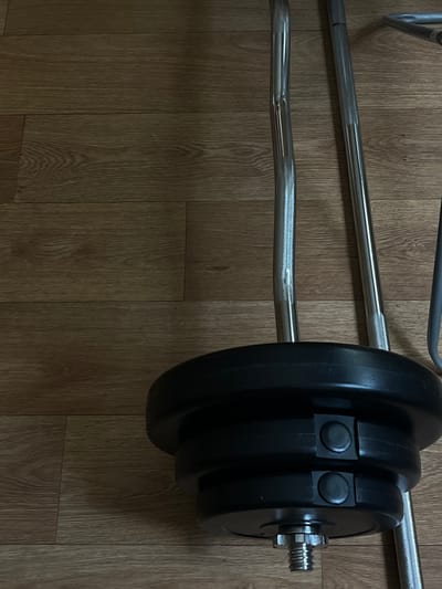 The image shows a close-up of weightlifting equipment on a wooden floor. In the foreground, a black weight plate with two circular black clamps is attached to the end of a barbell. The barbell, made of chrome-plated metal, has a knurled grip section. Behind this, another barbell, also chrome-plated, is visible. A third metallic bar, possibly part of a weight rack or another piece of equipment, is partially visible on the right edge of the frame. The wooden floor displays a wood grain pattern. There are no people present in the image. The scene depicts indoor exercise equipment in a domestic or gym setting. The specific location in Lviv, Ukraine, cannot be determined from the visual information provided.