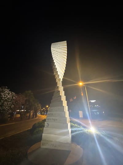 This is a nighttime, low-angle, full shot of a tall, spiral-shaped monument in the city of Bar, Montenegro. The monument is made of stacked white blocks, each inscribed with a place name and a distance in kilometers. The text is legible on many of the blocks, including "SOUTH POLE 14660km" at the bottom, "Leptis Magna 1132 km", "AKRA 4476km", "Etna 596km", "HILANDAR 436km", "JERUSALEM 1832km", "Petra 1960km", "MECCA 3000km", "Acropolis 604km", "ADA BOJANA 33km", "TEMPLE OF ZEUS 535km", "Victoria Lake 5095km", "EREBUS 15798km", and "VICTORIA FALLS 6003km".
To the left of the monument, there are blooming trees with white and pink flowers, and palm trees. Beyond the trees, a street with traffic lights and streetlights is visible. A car with its headlights on is parked on the side of the road. In the background, there is a building with illuminated signage that reads "HOTEL" and "PRINCESS".
To the right of the monument, a street lamp with a bright orange light illuminates the area, creating a strong lens flare effect that beams across the image. Two small orange flags are hanging from poles near the street lamp. The ground around the monument is covered with grass and low-lying bushes. The sky is dark and clear. The monument appears to be a significant landmark in the city.