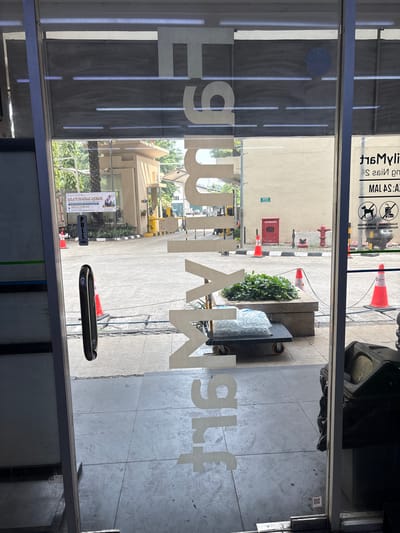 The image captures a view through a glass door, likely at the entrance of a commercial establishment. Prominently displayed on the glass are large, white, stylized letters that spell "TRUE MART" vertically. The letters are partially obscured by reflections from the environment outside.
The exterior scene visible through the glass shows a paved area, possibly a sidewalk or walkway, with some vegetation in planters. Orange traffic cones are placed strategically, one near a planter and another further in the background. A red fire hydrant is visible on the right side of the exterior, and a red utility box is mounted on a beige wall. Beyond this, a street scene with vehicles and buildings is implied, though not clearly detailed.
On the left side of the interior, near the glass door, a black door handle is visible. The floor inside appears to be tiled. On the far right, a black trash bin is present.
The text "MAL AS: A2" and "MALAYSIA" is visible on a sign on the exterior, suggesting the location might be related to Malaysia or have a Malaysian theme, which could be in North Jakarta, Indonesia, given the context provided. The overall impression is that of a retail space entrance, possibly a convenience store or a similar outlet.