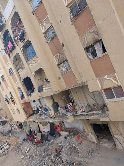 The image displays a multi-story residential building in Khan Yunis, Palestinian Territory, presenting extensive damage. The building's facade, predominantly beige with sections of faded orange, exhibits numerous cracks, impact points, and areas where plaster is missing. Many windows are shattered, with openings on various floors covered by black or white plastic sheeting, fabric, or blankets. Soot marks are visible around several window frames, particularly on the orange sections of the building. Laundry, including brightly colored clothes, hangs from multiple balconies, suggesting potential continued habitation or an attempt at normalcy. The lower levels of the structure are severely compromised, featuring fractured concrete slabs, exposed twisted rebar, and visible interior spaces. A significant amount of debris, including broken concrete, metal fragments, and various discarded items, litters the ground directly in front of and below the damaged sections of the building. Black plastic water tanks are observable on at least two lower-level balconies. The ground extending from the building is unpaved, covered in loose dirt and rubble. No individuals are discernible within the scene.