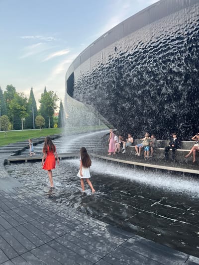 In this photograph taken in Krasnodar, Russia, several people are interacting with a water feature. The scene appears to be outdoors, with a bright sky and lush green trees visible in the background, suggesting it is daytime with pleasant weather.

The main focus is on a tiered water fountain where people are standing and playing. In the foreground, two young girls are visible. One girl wears a bright orange dress and is standing in the water, her feet splashing. To her left and slightly behind her, another girl in a white dress is also in the water, with her legs extended. Further in the background, a child in a light blue dress is standing in the water.

Behind these children, on a higher level of the fountain, a group of people are seated and standing. They are dressed in casual summer clothing, with some individuals holding what appear to be phones, possibly taking pictures or videos of the fountain or the surroundings. The water cascades down the different levels of the fountain, creating a dynamic and refreshing atmosphere. The architecture of the fountain is modern, with dark, sleek surfaces and a curved wall that seems to be part of a larger structure. The ground surrounding the fountain is paved with dark tiles. The overall mood of the image is joyful and relaxed, with people enjoying a sunny day by the water.
