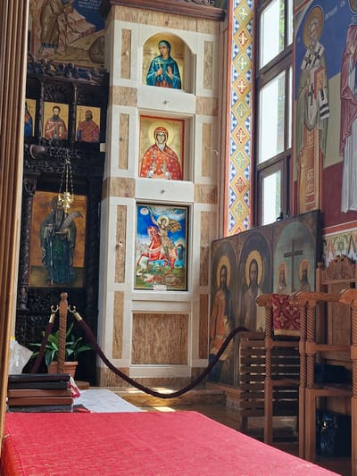This media file depicts the interior of a Christian Orthodox church in Sozopol, Bulgaria. The scene is dominated by religious icons and frescoes adorning the walls and ceiling. On the wall, several framed icons are visible, featuring depictions of saints and religious figures in traditional Byzantine style. One prominent icon shows Saint George on horseback battling a dragon. Above, the ceiling is covered in frescoes with similar religious imagery.
To the right, a wooden iconostasis, or icon screen, is visible, featuring multiple icons arranged in tiers. The wood is intricately carved and appears to be old. A red rope, possibly a barrier for visitors, is draped across part of the scene, connecting to decorative wooden posts. Further to the right, a large red fabric, possibly a liturgical vestment or altar cloth, is prominent. Natural light streams in from windows visible at the top of the frame, suggesting it is daytime. The overall atmosphere is one of reverence and historical significance, characteristic of an established religious site. The visible text on the icons is in Cyrillic script, consistent with the Orthodox tradition in Bulgaria.