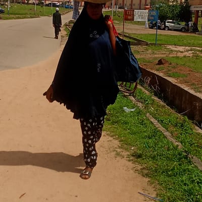 The image captures a sunlit street scene in Akwanga, Nigeria, featuring several individuals and elements of an urban environment. In the foreground, a woman walks along a sandy path bordering a paved road. She wears a dark blue or black flowing garment with white embroidered patterns, patterned trousers, and sandals. She carries a red-strapped bag over her shoulder and appears to have a wide-brimmed hat on her head, partially obscuring her face.
Further down the paved road, a man dressed in dark attire and a white skullcap walks away from the camera, casting a noticeable shadow, suggesting a bright, sunny day. The road appears to curve slightly. To the right, there's a strip of green grass and a concrete drainage channel.
In the mid-ground on the right side, a blue and white sign is prominently displayed, bearing the French text: "CERCLE FRANCAIS COE. AKWANGA SOYI LES BIEN VENUES". Beyond the sign, a silver car, a motorcycle, and several other people are visible near some buildings, indicating activity. The overall setting is an open-air public space, likely a street or pathway within a community.