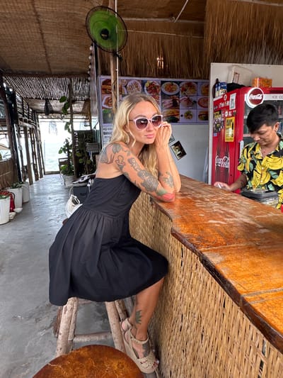 A woman with blonde hair and tattoos on her arms is sitting at a wooden table in what appears to be a beachside establishment. She is wearing sunglasses and a black dress. Beside her, a man in a colorful shirt with bananas on it is standing and looking down, possibly engaged in a transaction or conversation. Behind them is a red Coca-Cola refrigerator and a display of food menu pictures. The setting is rustic, with thatched roofing and woven bamboo furniture. A green fan is mounted on a wooden post. In the background, there is a view of the water and more wooden structures. The overall ambiance suggests a casual, tropical location. Visible text includes "Coca-Cola". The time of day appears to be daytime.