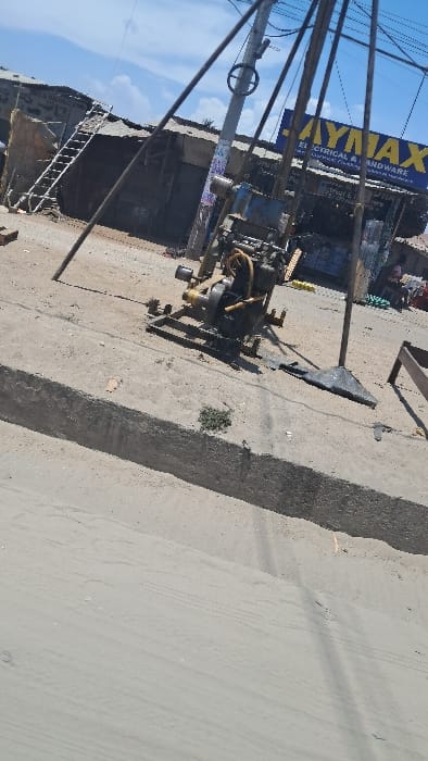 The scene depicts an outdoor urban environment in Shimo La Tewa ward, Kenya, under a clear blue sky with scattered white clouds. In the midground, a portable borewell drilling rig is positioned on a dirt and gravel surface. The rig consists of a yellow-painted engine unit, likely a diesel engine, mounted on a steel frame, with multiple black metal poles forming a tripod-like support structure. A black, triangular metal base plate lies on the ground to the rig's right. Directly behind the rig stands a utility pole with a circular attachment and overhead wires. The immediate foreground shows a light-colored, sandy or dirt surface, separated from the midground by a low, raised concrete barrier or curb. Subtle tire tracks are visible on the sandy surface. In the background, a row of single-story commercial buildings with corrugated metal roofs is present. One building features a prominent blue and yellow sign displaying "JAYMAX ELECTRICAL & HARDWARE." An aluminum extension ladder leans against the front of an adjacent building to the left. Indistinct human figures are visible near the commercial structures in the far background. No specific actions or interactions are clearly discernible.