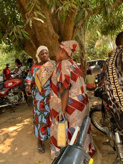 This image captures an outdoor scene in Akwanga, Nigeria, under bright daylight with dappled shadows. The ground is unpaved dirt. In the mid-ground, a large tree with a thick, textured trunk and green foliage provides overhead canopy. The central focus is on two women engaged in conversation. The woman on the left, facing towards the right, wears a light blue dress adorned with red, green, and yellow patterns, a white headwrap, and a cream-colored patterned shawl draped over her left shoulder. She has a silver hoop earring in her visible left ear and wears blue flip-flops. Her right hand holds a small, dark, indistinct object. The woman on the right, facing towards the left, wears a brown, red, and cream patterned dress with a matching headwrap. She has a prominent gold hoop earring in her visible left ear and carries a rectangular, light-colored (possibly yellow or beige) bag with gray handles in her left hand. Her right side is partially obscured by the black handlebar of a motorcycle in the extreme foreground.
Behind these two women, several other individuals and vehicles are present. To the far left, a person wearing a red shirt and a dark cap stands near a red motorcycle. In the middle-right background, an individual wearing a light green top and a reddish-orange wrap is seated on a low object. Further back, a dark-colored utility vehicle or van is parked, with additional people visible inside and around it. The back of a person wearing a black, white, and gold patterned shirt is visible in the right foreground. The background is characterized by dense green foliage, including palm-like fronds, indicative of a tropical environment.