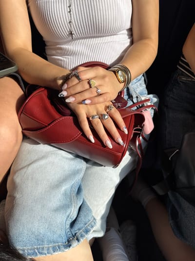 A female subject, located in the city of Getap, Armenia, is depicted seated, wearing a white ribbed tank top and light blue denim shorts. A silver chain with a cross pendant hangs on her chest. Her hands are clasped, resting on a red structured handbag. Multiple silver-toned rings adorn her fingers, including a heart-shaped ring, a star-design ring, and a ring with an engraved heart. Her nails are long, almond-shaped, and decorated with glitter, blue tips, black polka dots, and small gems. A multi-strand silver-toned bracelet and a gold-toned watch are present on her left wrist. Direct sunlight illuminates her right arm and the handbag. A second individual is partially visible to the right, showing a leg in dark denim shorts, a striped garment, and a foot wearing a white sock and a white shoe. The immediate environment suggests an enclosed space, with a dark background.